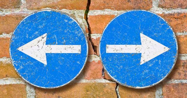 Durch eine Backsteinmauer, als Symbol eines Hauses, verläuft fast senkrecht ein Riss, rechts und links davon befinden sich zwei blaue Schilder mit weißen Pfeilen, die jeweils nach rechts und links, also vom Riss weg, zeigen, als Symbol für das Scheidungspaar, dass getrennte Wege geht | Scheidungsimmobilie