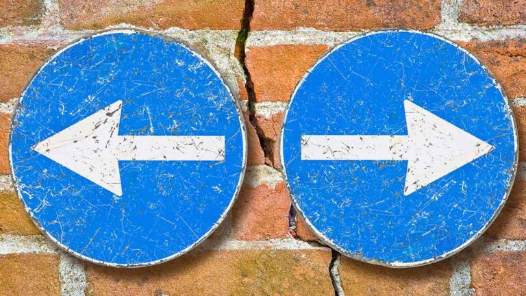 Scheidungsimmobilie Durch eine Backsteinmauer, als Symbol eines Hauses, verläuft fast senkrecht ein Riss, rechts und links davon befinden sich zwei blaue Schilder mit weißen Pfeilen, die jeweils nach rechts und links, also vom Riss weg, zeigen, als Symbol für das Scheidungspaar, dass getrennte Wege geht | Scheidungsimmobilie